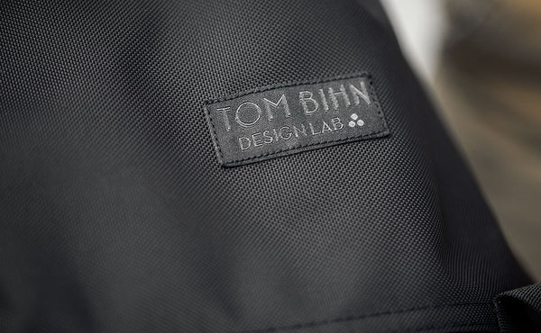 The Shadow Guide has the TOM BIHN Design Lab label; the label is rectangular, smaller than our original label, and has a black background with dark grey text in the foreground. The three dots are the Therefore sign.